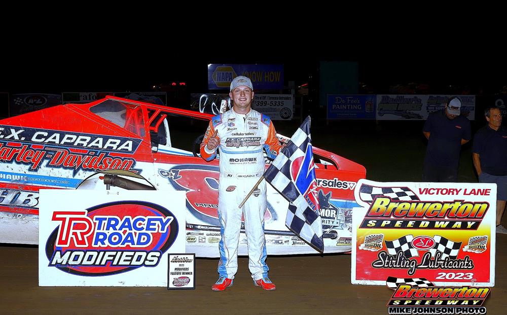 Race car driver in a white jumpsuit stands next to a red and white race car holding a checkered flag at Brewerton Speedway. Signs around him read 'Tracey Road Modifieds' and 'Brewerton Speedway Victory Lane
