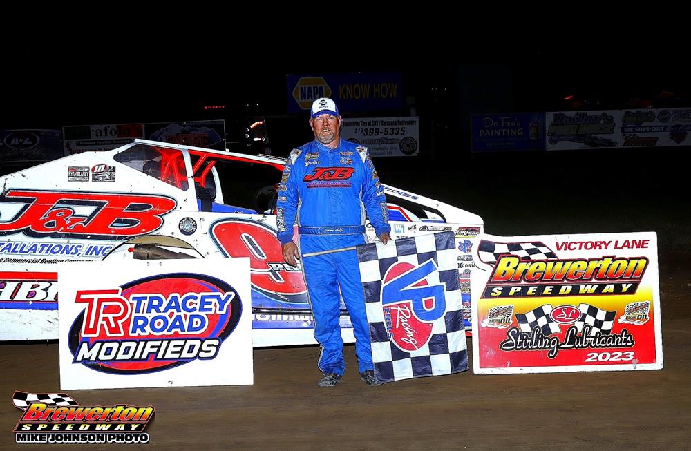 Race car driver in a blue jumpsuit stands next to a white race car and two large signs at Brewerton Speedway. One sign reads 'Tracey Road Modifieds' and the other 'Brewerton Speedway Victory Lane'. The driver is holding a checkered flag.