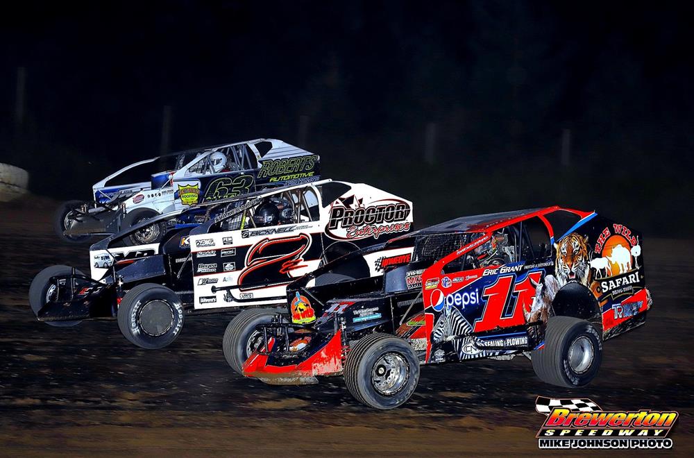 Three modified race cars competing on a dirt track at Brewerton Speedway under the night sky. The cars are brightly colored with sponsorship logos visible, and dirt is flying as they speed around the track.