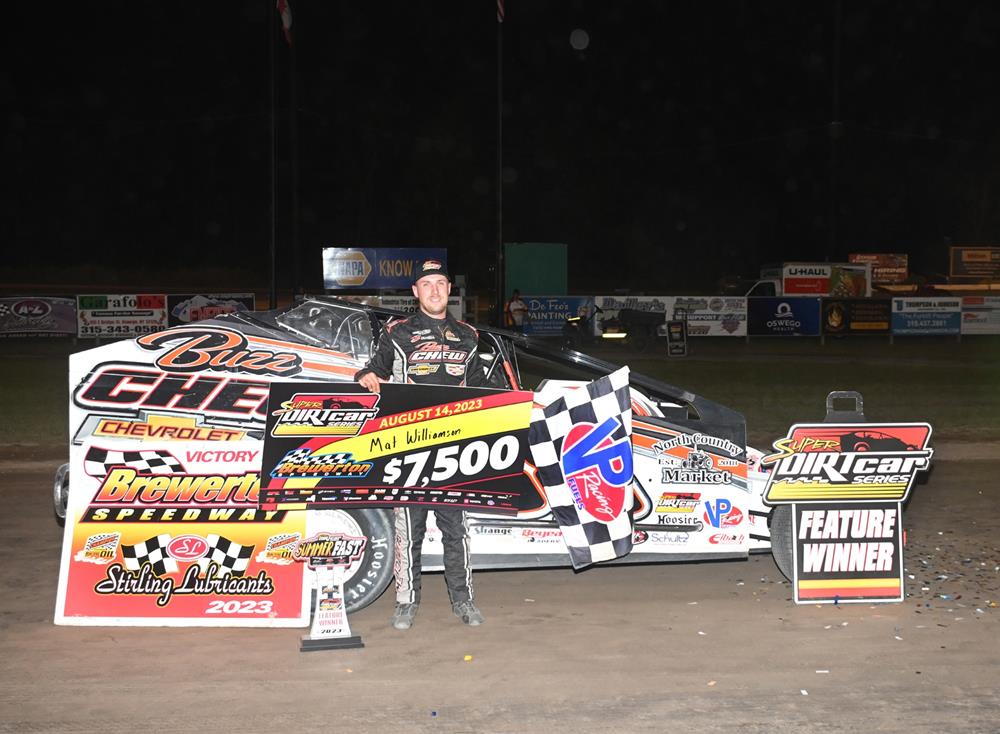 Race car driver in a black jumpsuit stands next to a black and white race car holding a large check for $7,500 and a checkered flag at Brewerton Speedway. Signs around him read 'Brewerton Speedway Victory Lane', 'Super DIRTcar Series', and 'Feature Winner'.