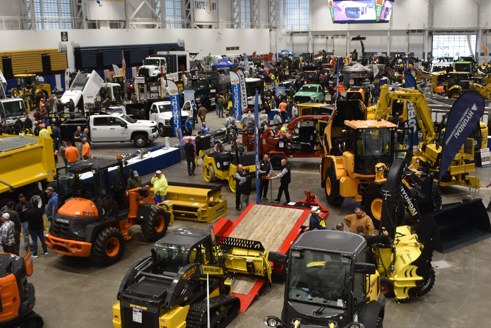 An indoor exhibition hall filled with various construction equipment and machinery, including loaders, excavators, and trucks. Crowds of people are walking around, examining the equipment and engaging with exhibitors.