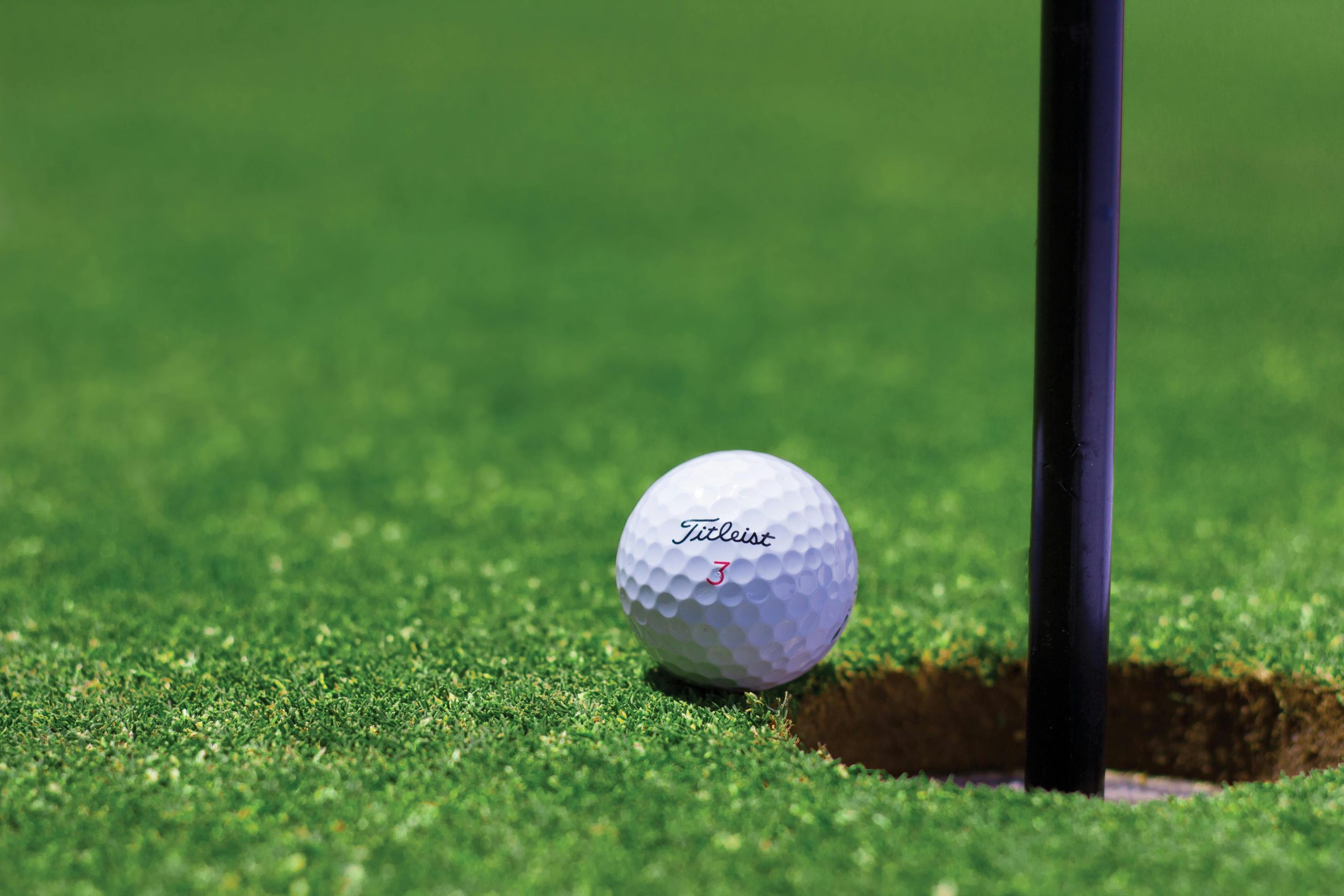 A close-up shot of a Titleist golf ball positioned near the edge of a hole on a lush green putting surface.