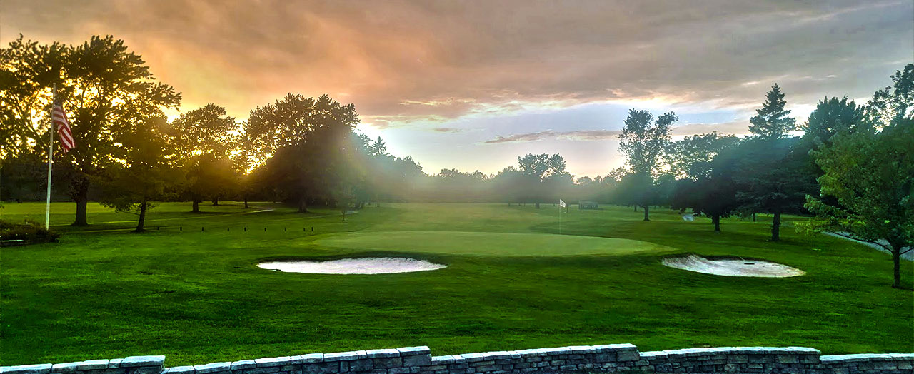 Sunset view of a golf course with a green field, sand bunkers, and trees in the background. An American flag is visible on the left side of the image.