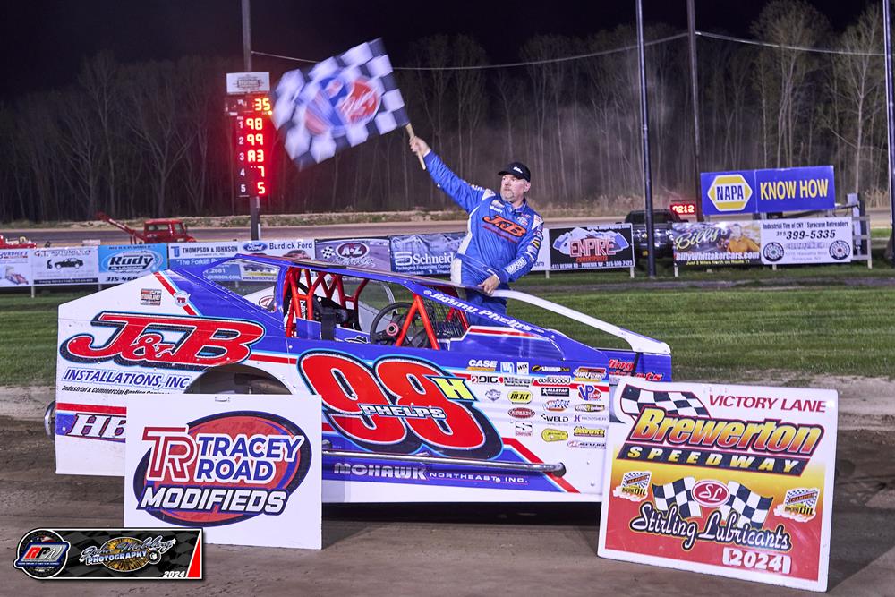 Race car driver celebrating atop a blue and red car in Victory Lane at Brewerton Speedway, holding a checkered flag, with Tracey Road Modifieds and Brewerton Speedway signage.