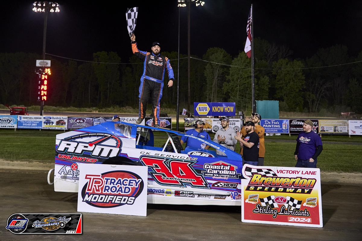 Race car driver celebrating atop a blue car in Victory Lane at Brewerton Speedway, holding a checkered flag, with Tracey Road Modifieds and Brewerton Speedway signage.