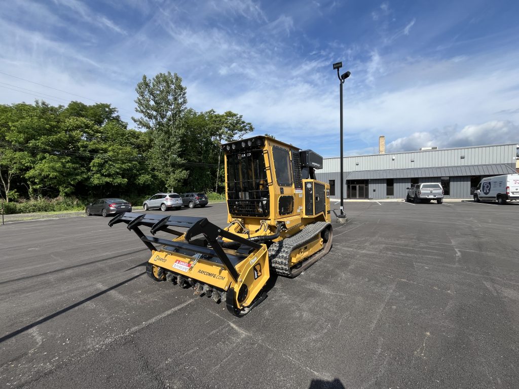 Rear side view of a yellow Rayco forestry mulcher on a paved lot with trees, cars, and a building in the background.