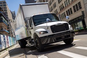 Freightliner M2 106 box truck in white driving through a city street, front-side view.